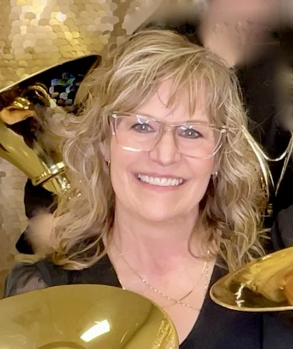 Smiling woman with wavy hair and glasses, Sherri Seward holds a brass instrument against a shiny gold sequin background.