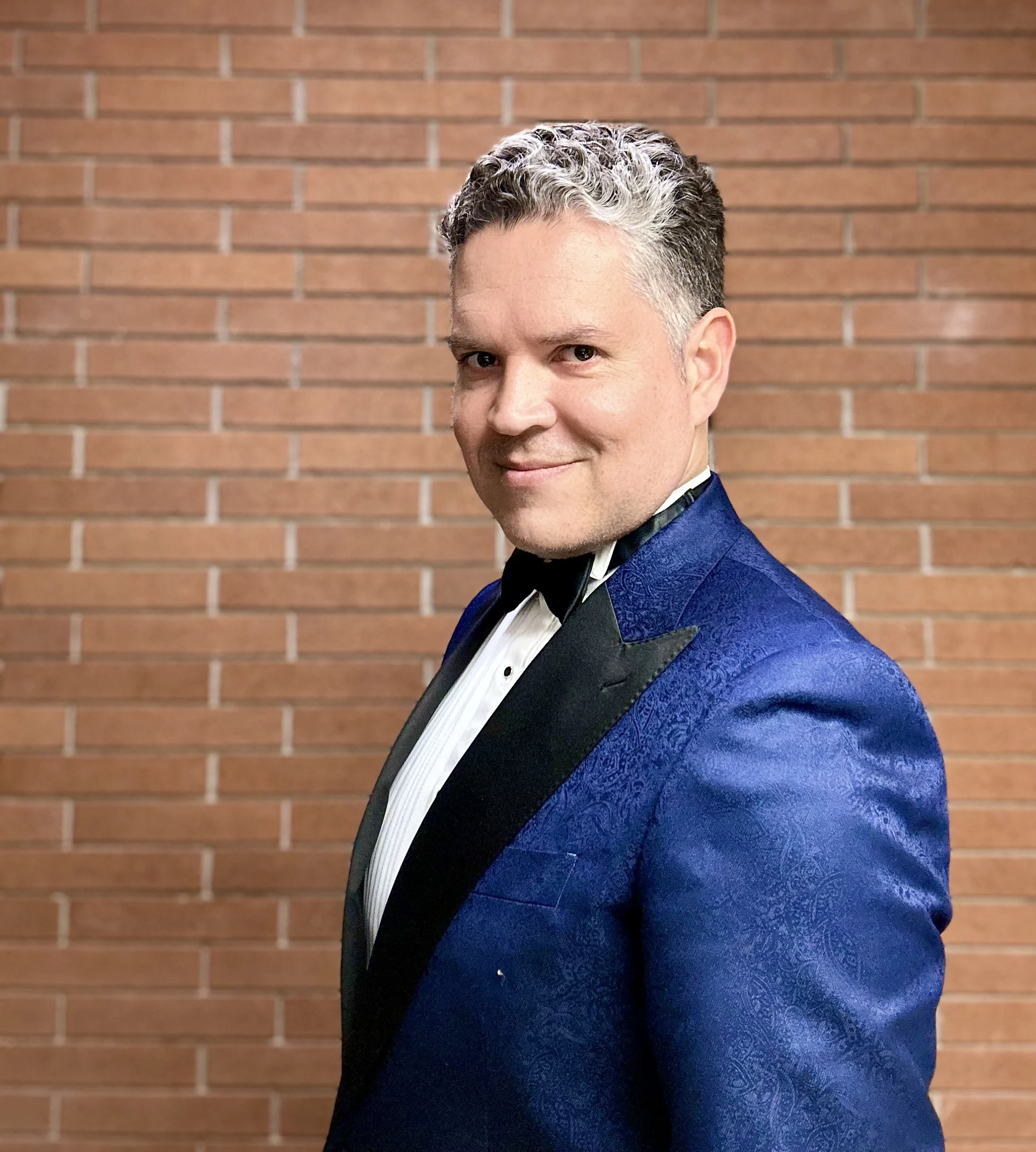 Diego Piedra, dressed in a blue tuxedo and bow tie, stands smiling at the camera in front of a brick wall.