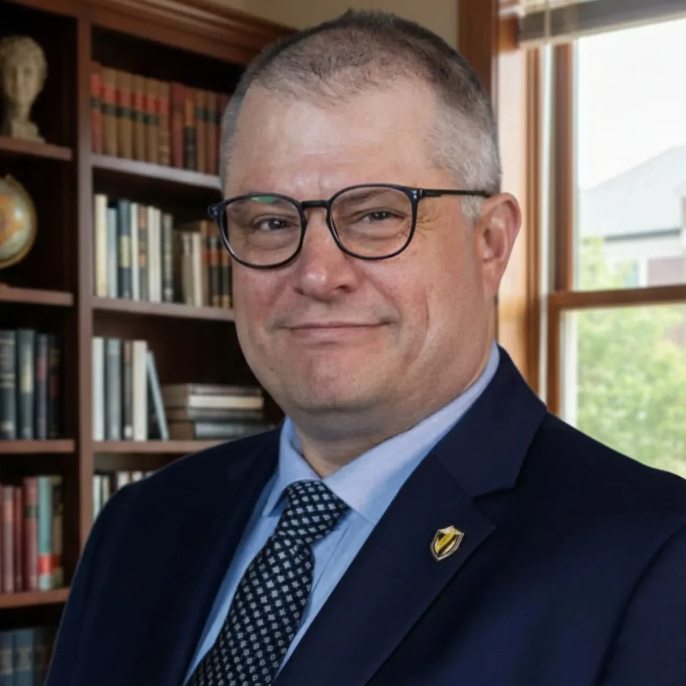A man in a navy suit and glasses stands in a library with shelves of books and a window in the background.
