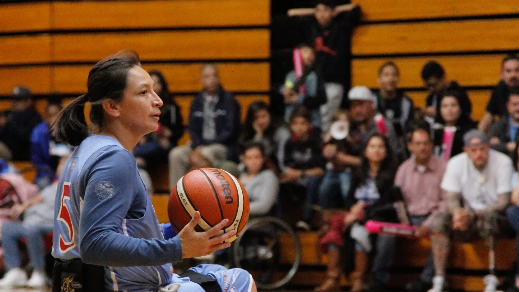 A woman athlete in a wheelchair holds a basketball on an indoor court, with a crowd watching in the background.