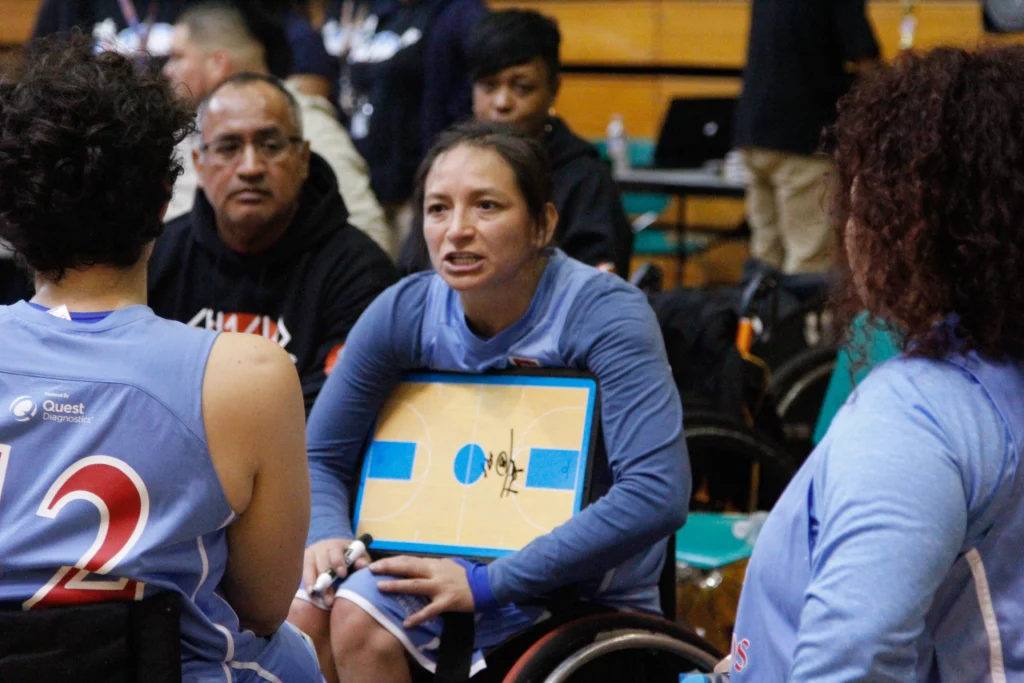 A woman athlete in a wheelchair holds a basketball tactics board, talking to teammates during a game.