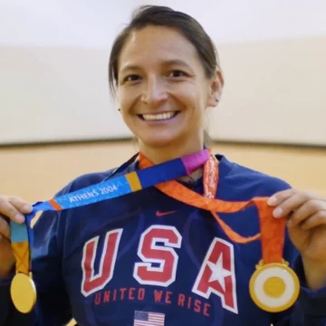 Smiling athlete and advocate wearing a USA shirt holds up two gold medals, one labeled "Athens 2004.