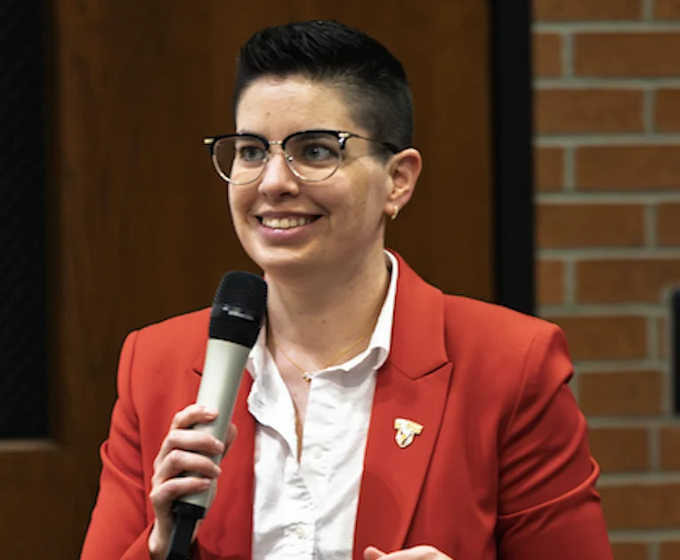 Stefanie Heinrich, a person with short dark hair and glasses, wears a red blazer while speaking into a microphone.