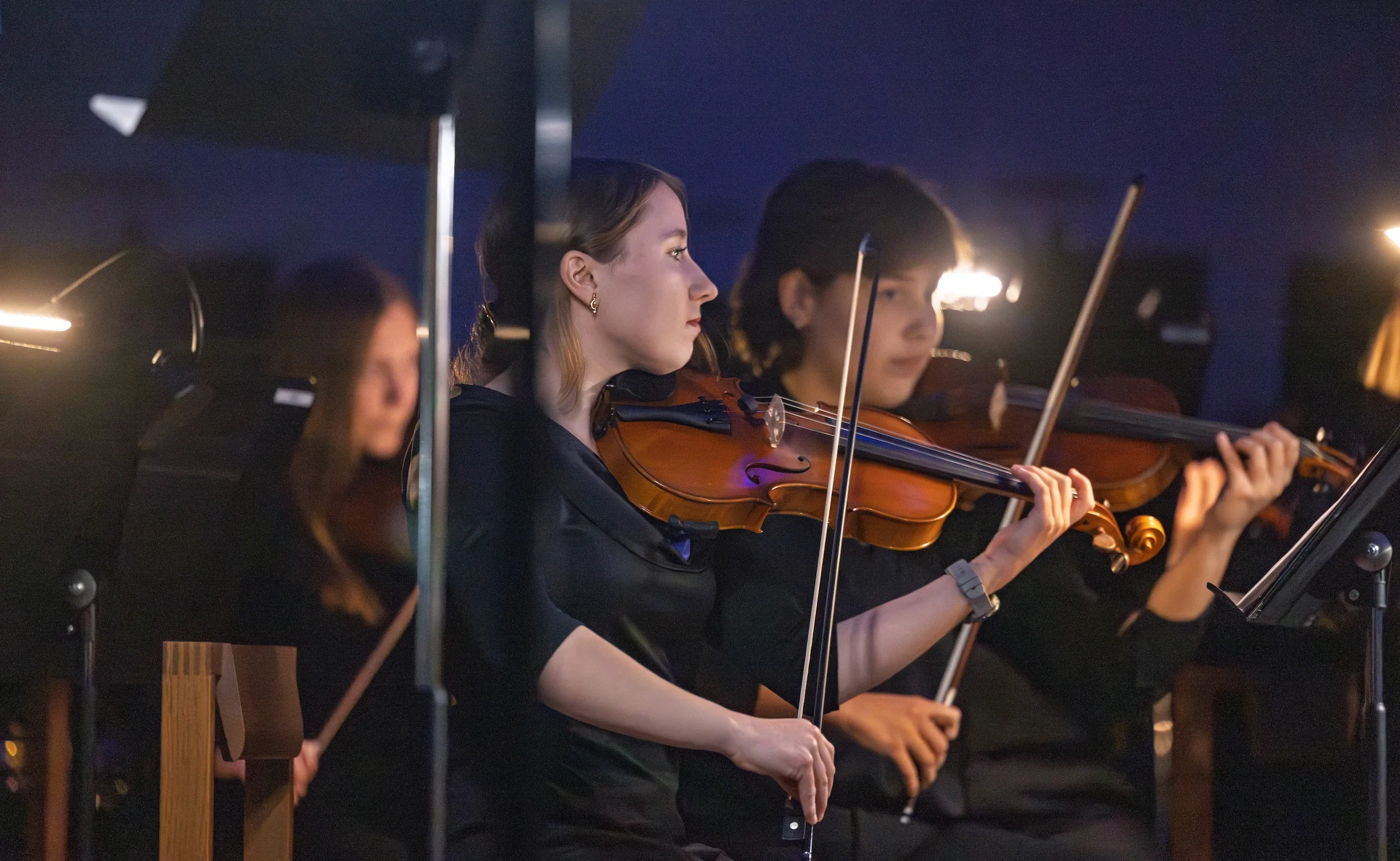 Two Valparaiso University students in black outfits play violins in an orchestra, immersed in music and illuminated by soft music stand lights.