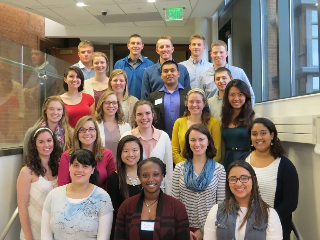 A group of twenty young adults, participants in the CAPS Fellows Program, smiling and posing together on an indoor staircase.