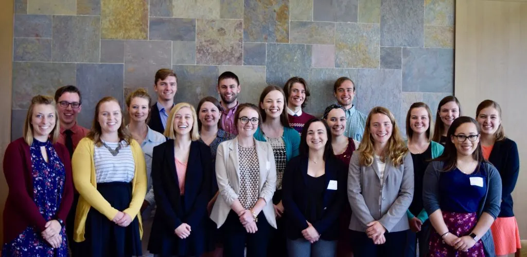 A group of 18 people, participants in the CAPS Fellows Program, stand smiling in front of a stone-tiled wall, dressed in business casual attire.