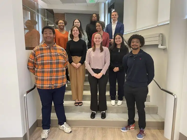Ten people stand on indoor stairs, smiling and posing together in casual attire as part of the CAPS Fellows Program group photo.