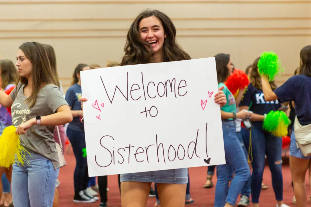 Smiling young woman holds “Welcome to Sisterhood!” sign at an indoor Valparaiso University Fraternity and Sorority Life event, surrounded by people and pom-poms.