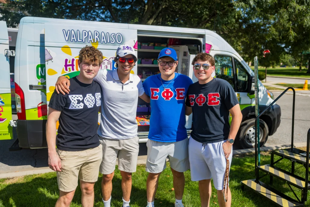 Four young men in Fraternity and Sorority Life shirts stand smiling in front of an ice cream truck on a sunny Valparaiso University day.