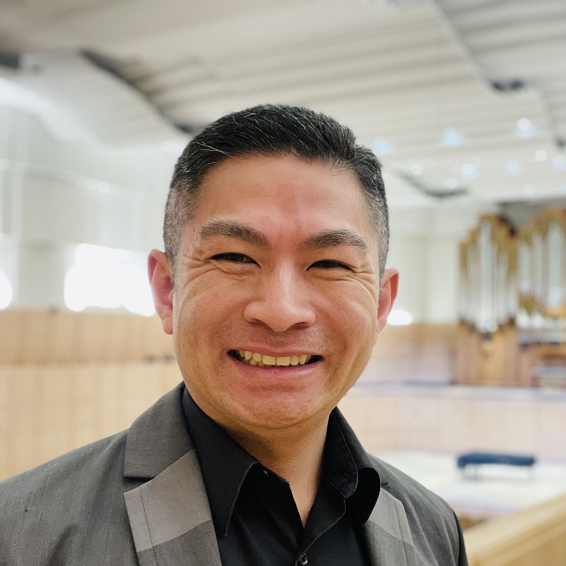 Smiling man in a gray suit stands indoors with a blurred concert hall and pipe organ in the background, identified as Haruhito Miyagi, Ph.D.