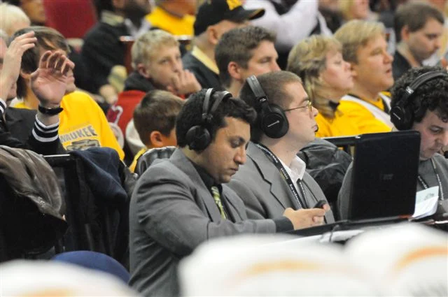 Sports announcers with headsets, including Adam Amin, sit at a table as fans watch a game in the crowded stadium background—Follow the Beacon for all the action.