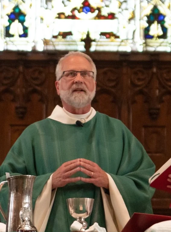 A priest in green vestments stands at an altar, hands together, with stained glass in the background, preparing for upcoming workshops within the church community.