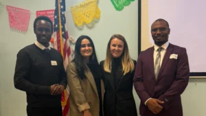 Four people in business attire stand smiling together indoors on the Valpo campus, with colorful banners and a US flag behind them.