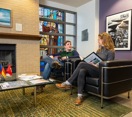 Two people sit and talk in a lounge with art, flags, a table of papers, and a stained glass window during a Faculty Workshop.