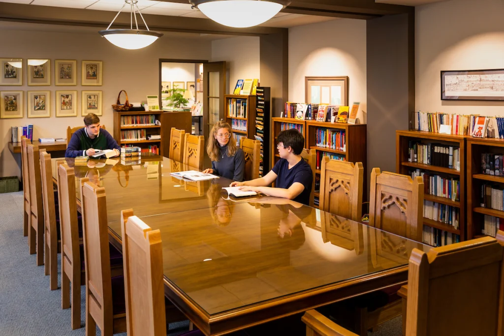 Three students study at a large wooden table in a quiet, well-lit library with bookshelves in the background, preparing for an upcoming Faculty Workshop.