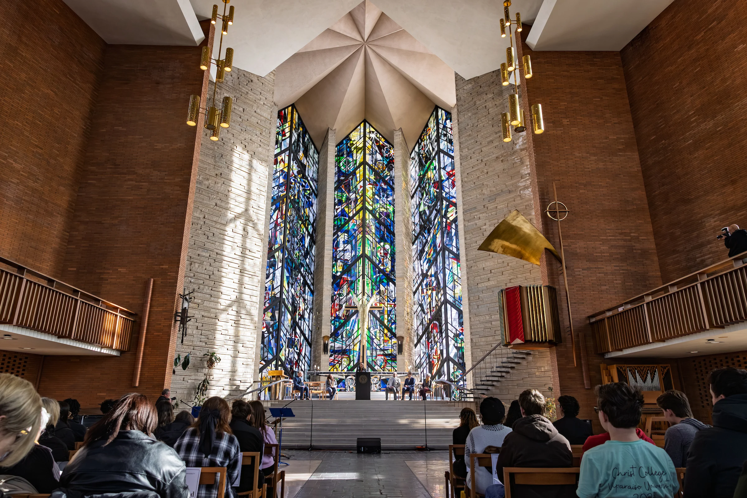 People sit in a modern church, grounded in faith, with tall, colorful stained glass windows behind the altar.