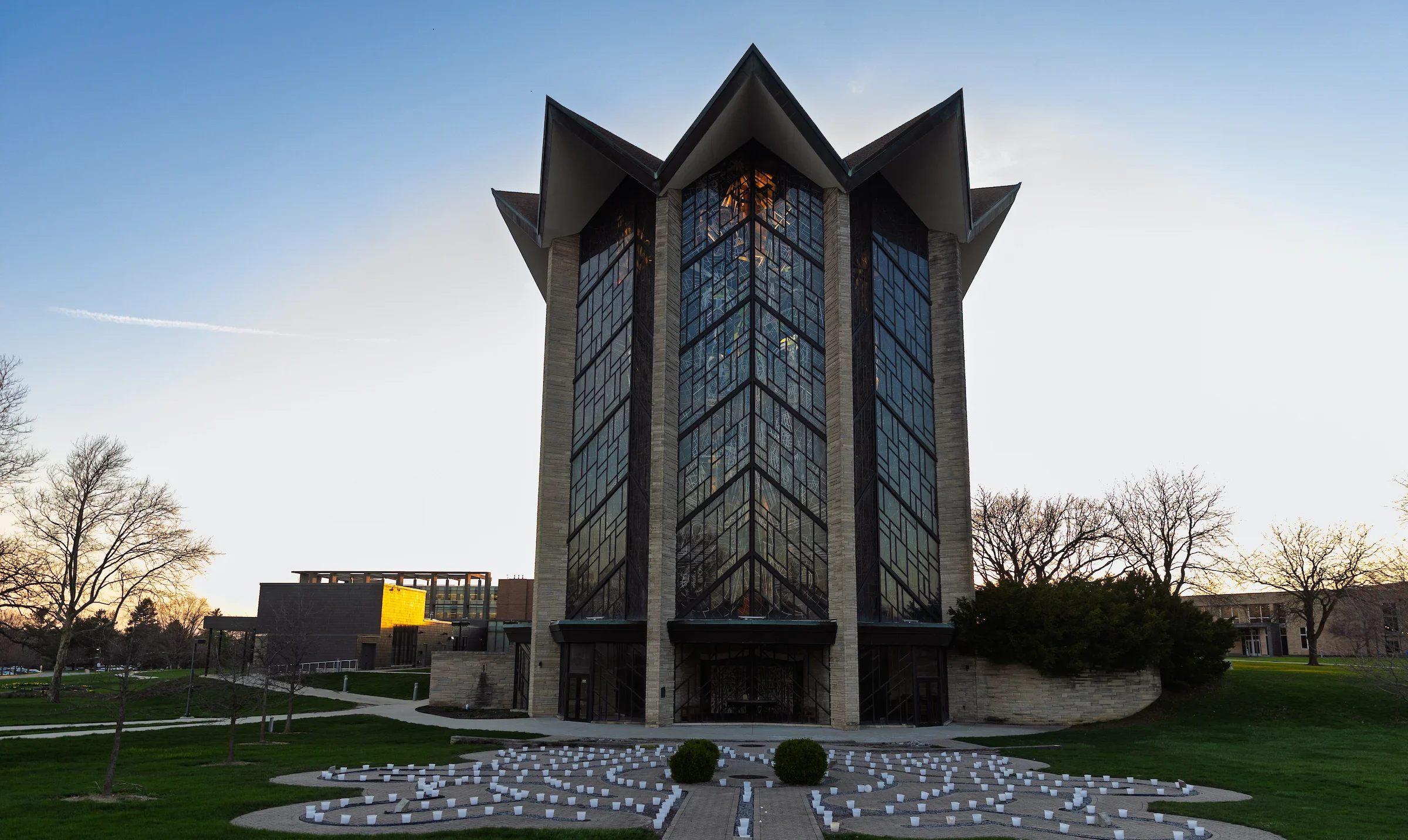 Modern glass chapel with sharp roof angles, at sunset, lights arranged on the grass in front—an inspired vision by Jackson Price, grounded in faith and modern design.