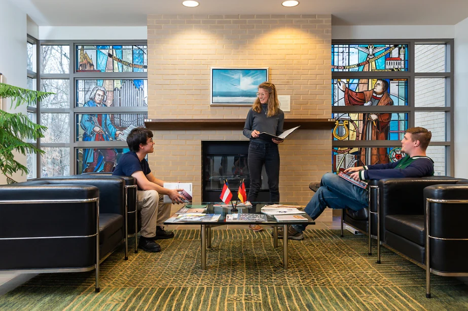 Three people talk in a modern lounge during a Faculty Workshop, with stained glass windows and a brick fireplace in the background.