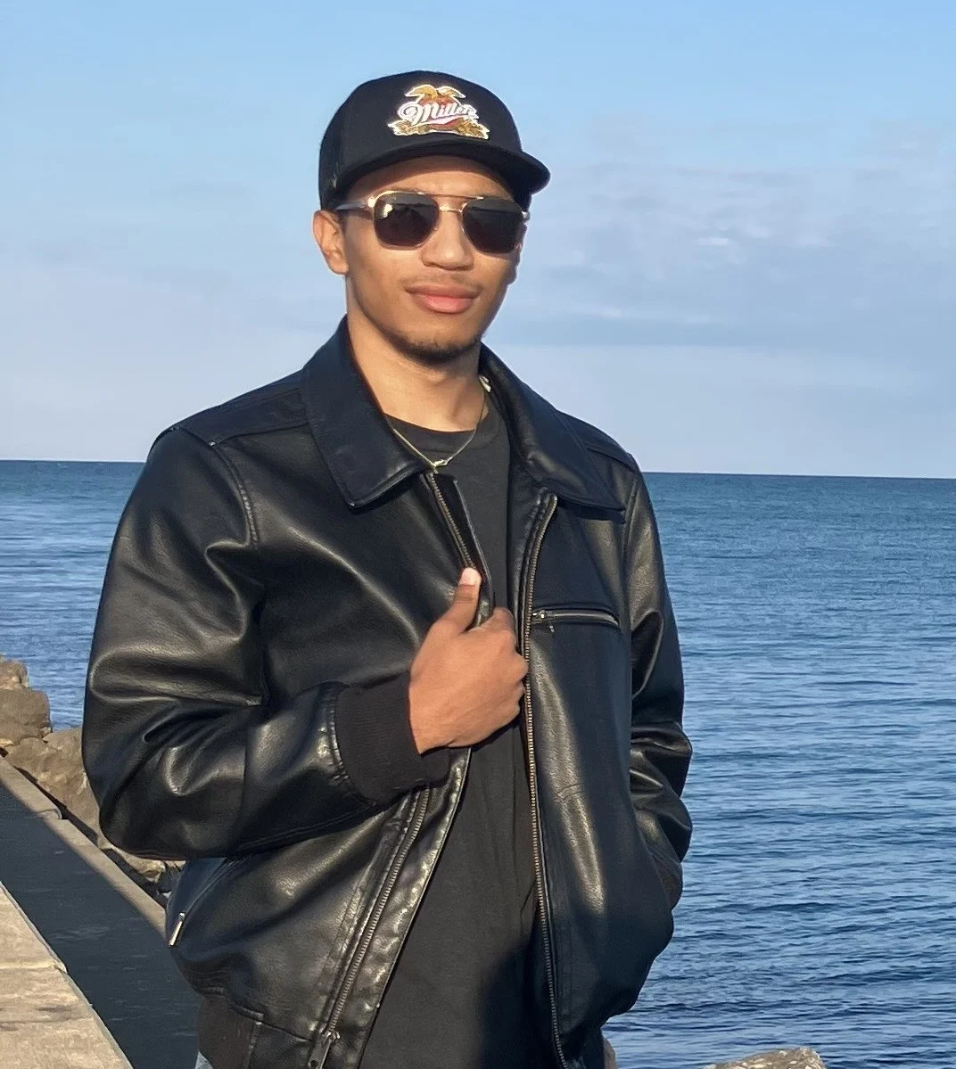 Young man in a black leather jacket and sunglasses stands by a body of water on a sunny day, capturing an effortless OMP Spotlight moment.