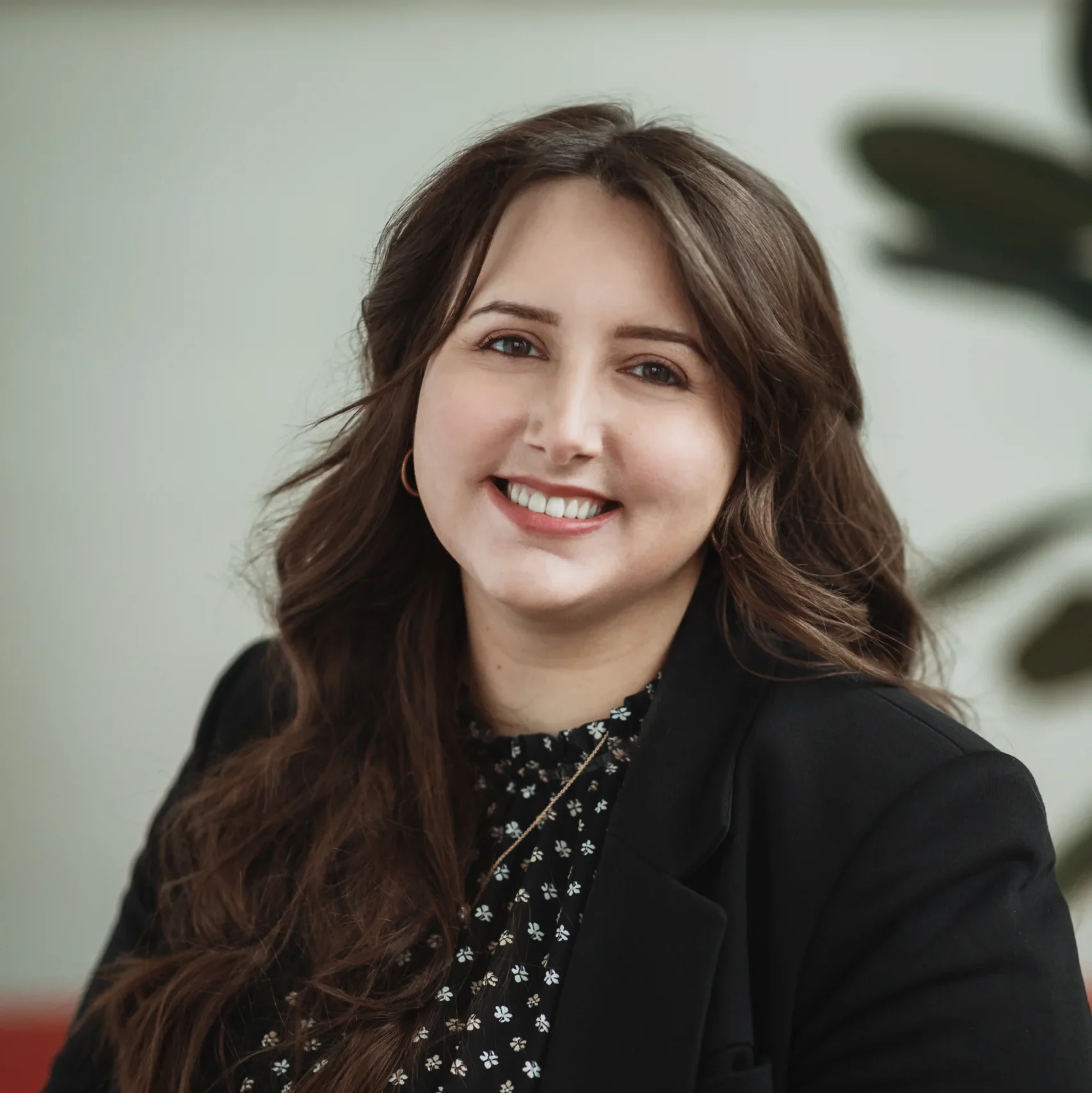 Woman with long brown hair smiling, Daniela Trajceski is wearing a black blazer and patterned top, with a blurred plant in the background.