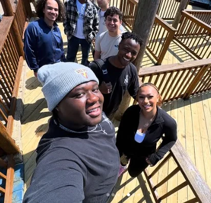 Six people smile and pose for an OMP Spotlight selfie on a sunny wooden deck with railings.