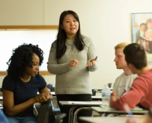 A woman stands and speaks in a classroom while three students sit and listen attentively around her, as she addresses the question: Is Mental Health a Public Health Issue?.