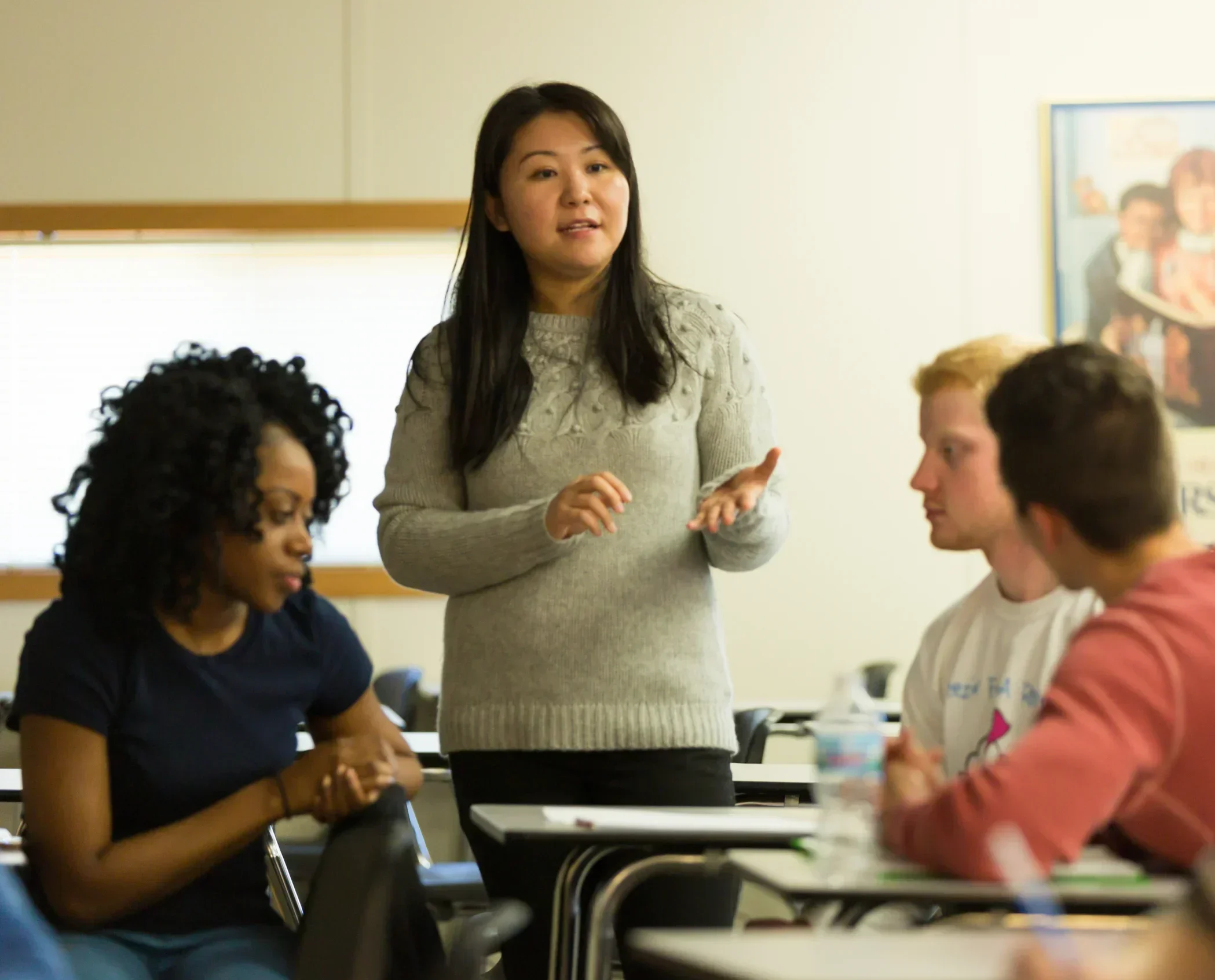 A woman stands and speaks in a classroom while three students sit and listen attentively around her, as she addresses the question: Is Mental Health a Public Health Issue?.