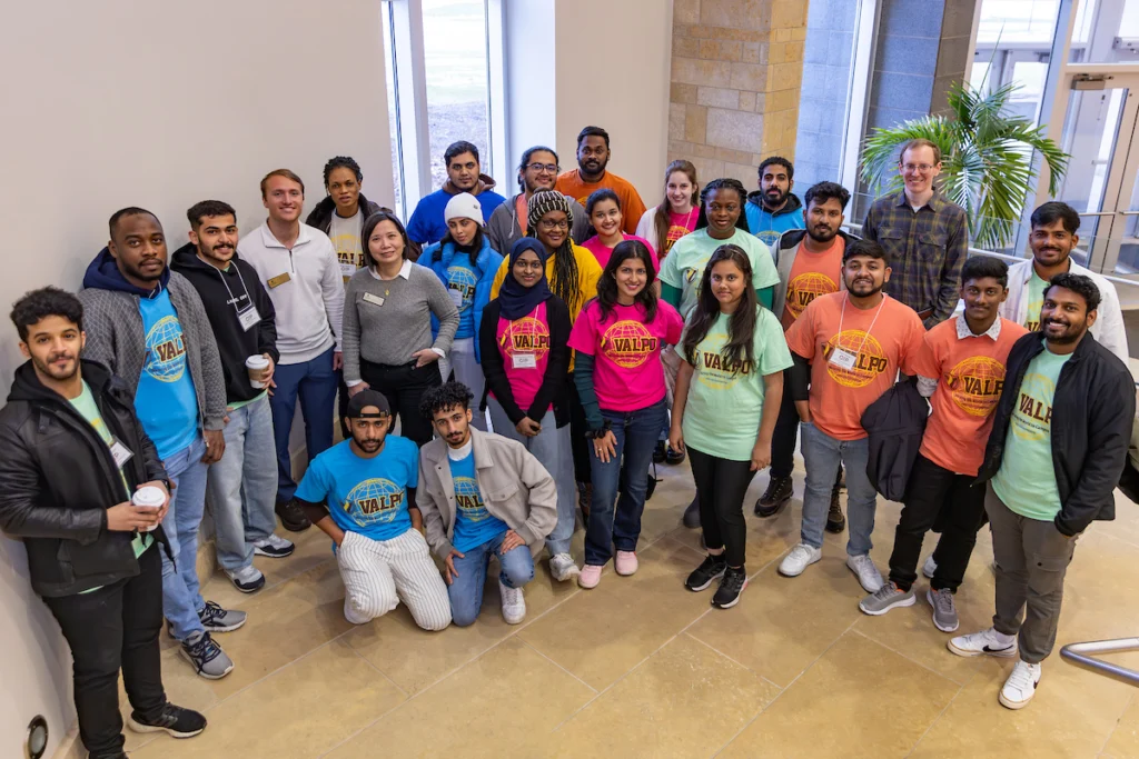 A diverse group of people in colorful "Valpo" shirts, representing International Academics, pose together indoors, smiling at the camera.