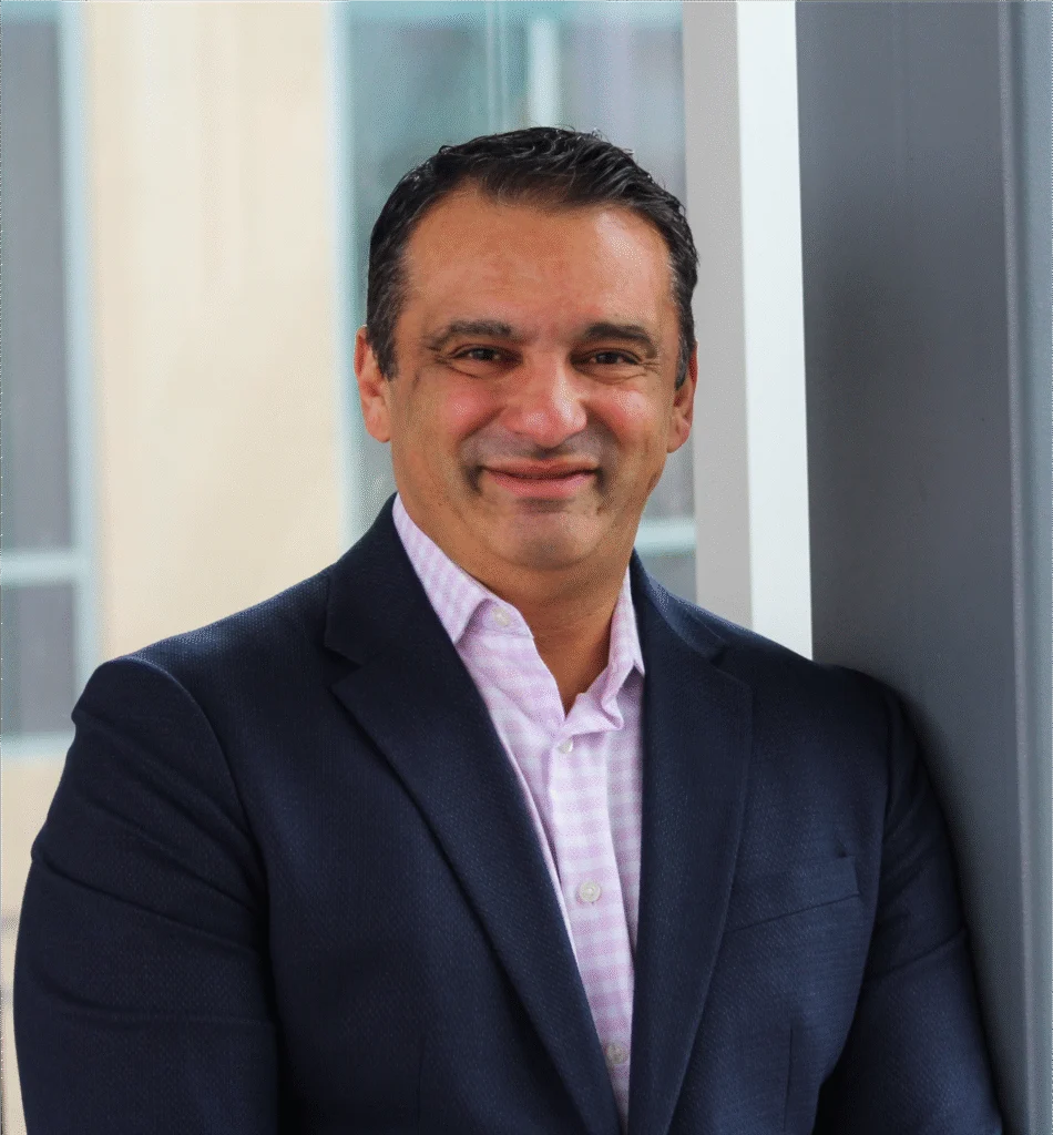 Man in a navy blazer and light pink shirt smiling, standing indoors by a window and gray wall at an Innovation Hub.