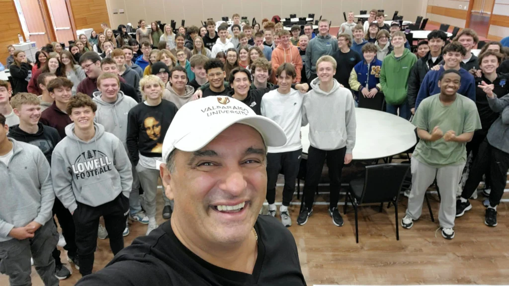 Man in a white cap taking a selfie with a large group of smiling students at the Innovation Hub classroom or event hall.