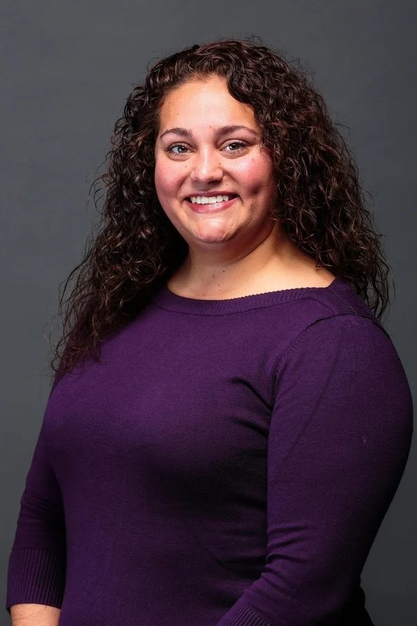 Smiling woman with curly brown hair wearing a purple sweater, posing confidently against a plain gray background—perfect for representing an Innovation Hub's creative spirit.