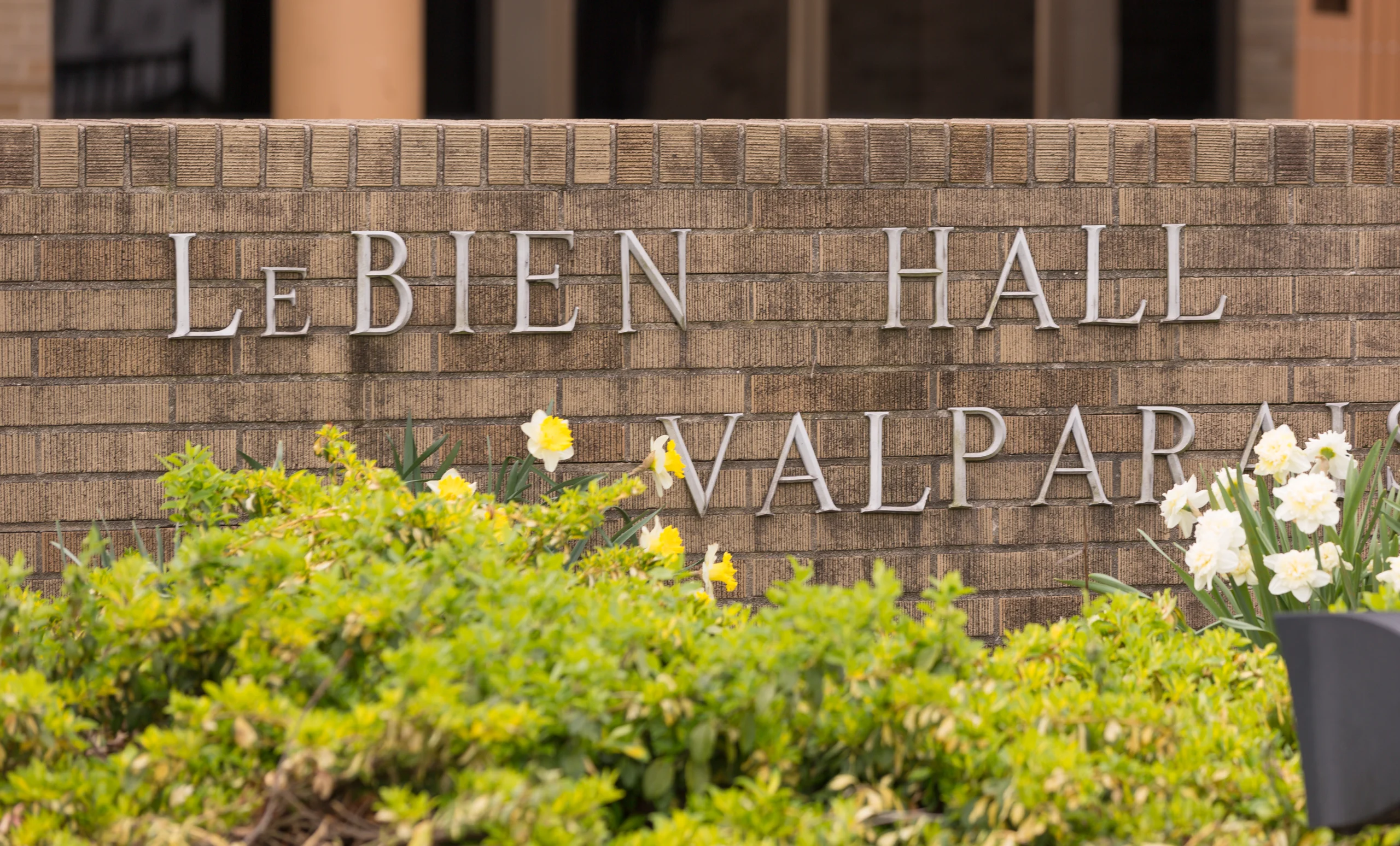 A brick wall sign reads "LeBien Hall Valparaiso," with yellow flowers and greenery in front—serving as a beacon for students and staff, including dedicated School Nurse Juanita Fox Gryfinski.