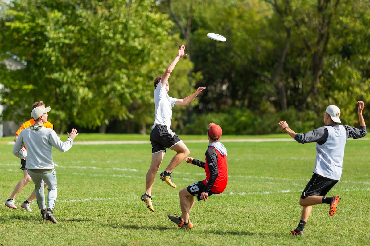 Four people play ultimate frisbee on a grassy field; one jumps to catch a frisbee in mid-air, showcasing agility and teamwork often seen in both traditional sports and esports.