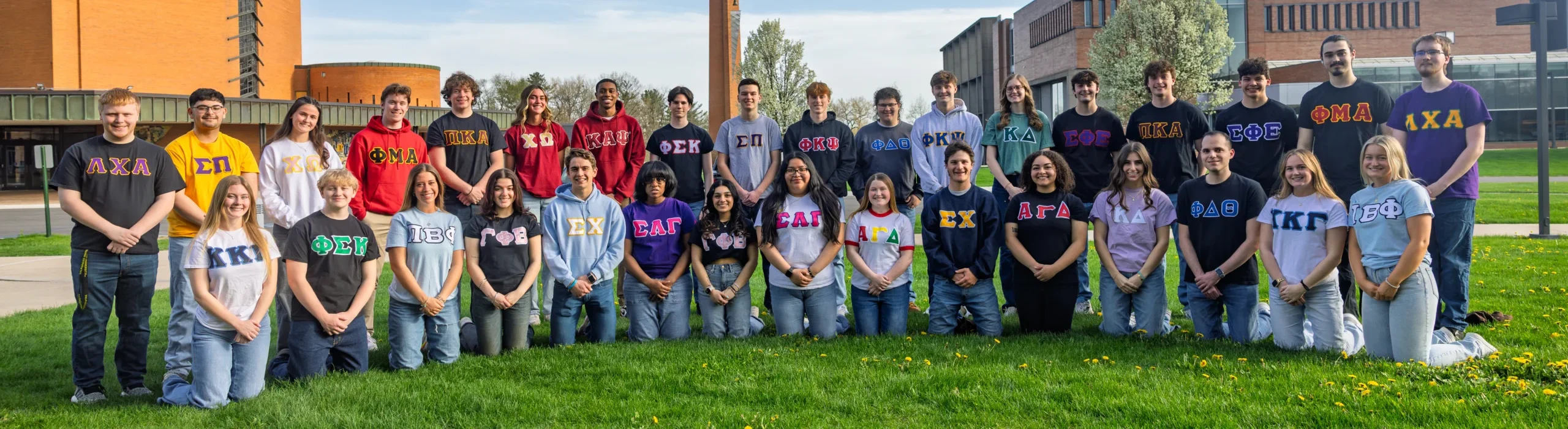 A group of students in Greek letter shirts pose together outside on a grassy area in front of campus buildings, capturing the spirit of Fraternity Life.