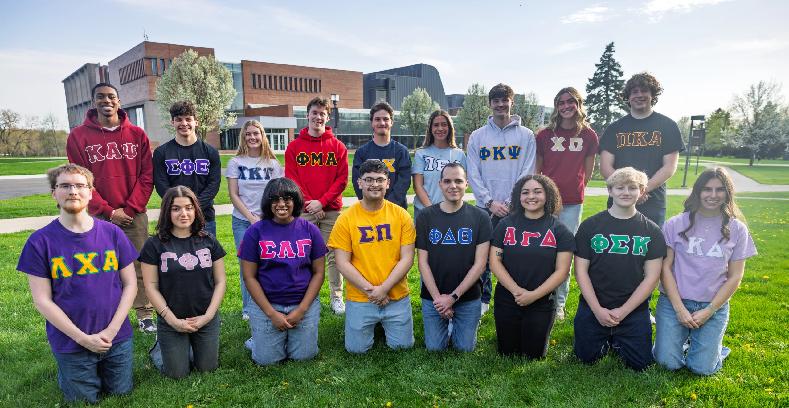 A group of students wearing colorful Greek letter shirts pose together on a grassy lawn outside, capturing the vibrant spirit of Fraternity Life.
