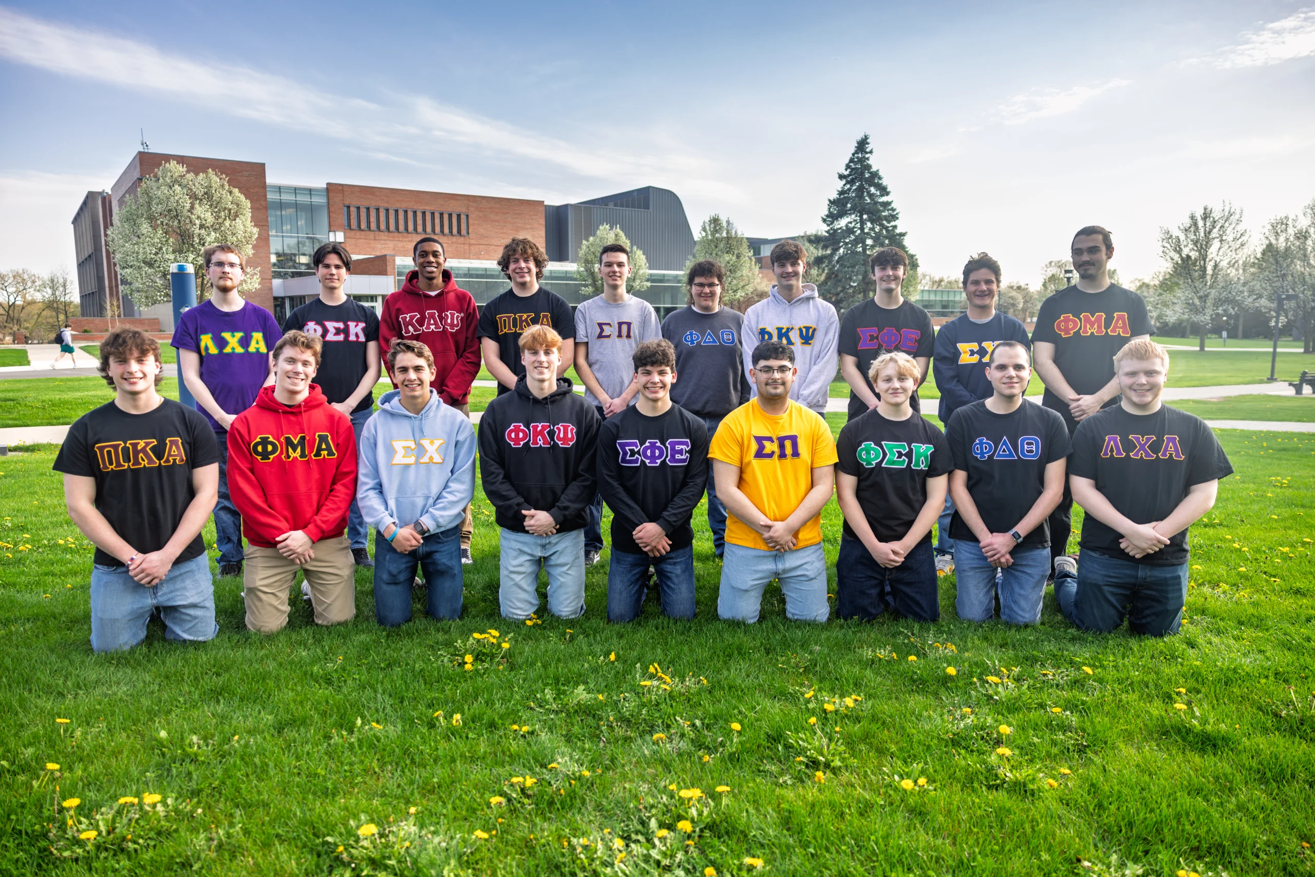 A group of students in fraternity shirts pose together on a grassy lawn in front of a campus building, capturing a spirited moment of Fraternity Life.