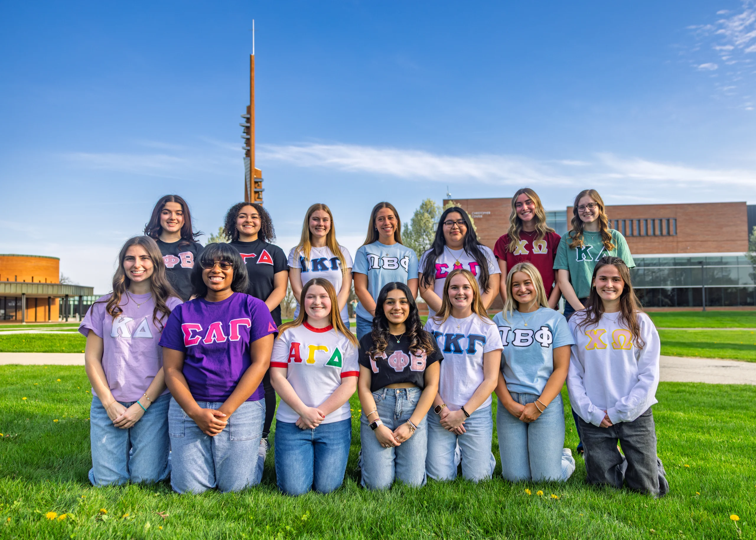 Thirteen women kneel on grass, wearing different sorority shirts with campus buildings in the background, capturing the spirit of Fraternity Life.