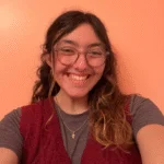 Smiling person with glasses and wavy hair in front of a peach-colored wall, wearing a red vest and gray shirt—one of our friendly Undergraduate Writing Consultants.