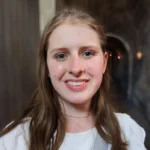 Woman with long brown hair smiling, standing in a dimly lit stone hallway, wearing a white top—one of our talented Undergraduate Writing Consultants ready to help you.