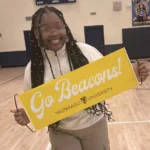 Smiling person holding a yellow "Go Beacons! Valparaiso University" sign in a gymnasium, celebrating with the Undergraduate Writing Consultants.