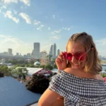 Smiling woman in red sunglasses with city skyline, Ferris wheel, and Undergraduate Writing Consultants enjoying the sunny day in the background.