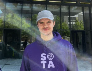 A person in a purple hoodie and gray cap stands outside the LINC Language and Intercultural Learning Center, sunlight creating a rainbow glare across the image.