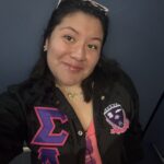 Young woman in a sorority jacket smiles softly at the camera with her head tilted to the side.