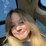 A young woman with blonde hair, one of our Undergraduate Writing Consultants, smiles inside a car with sunlight coming through the sunroof.