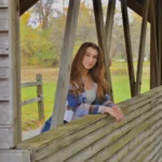 Young woman with long hair leans on a wooden railing in a rustic covered bridge, surrounded by autumn trees, reflecting the thoughtful presence of Undergraduate Writing Consultants.
