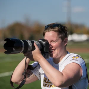 Person smiling while taking a photo with a large camera lens outdoors on a sunny day, capturing the perfect shot inspired by Beacon's Reflection.