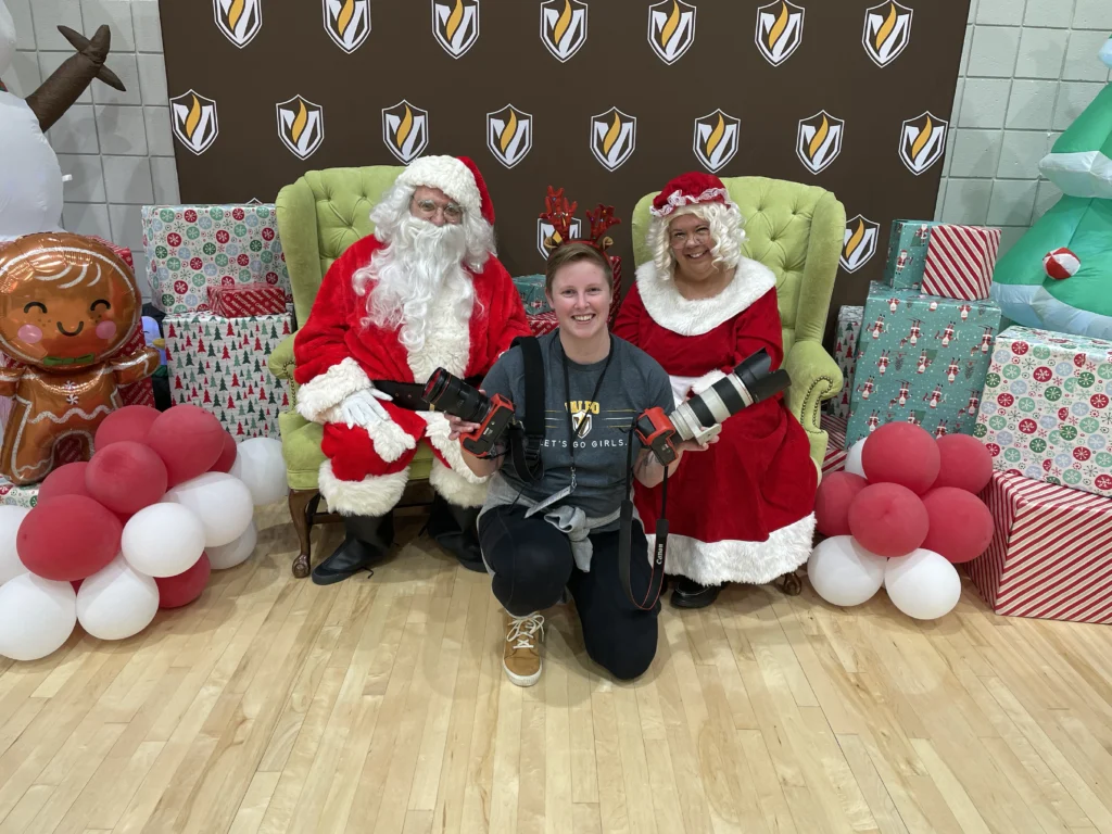 A smiling woman kneels with cameras in front of Santa and Mrs. Claus, surrounded by gifts and holiday decor—a festive moment captured for Beacon's Reflection.