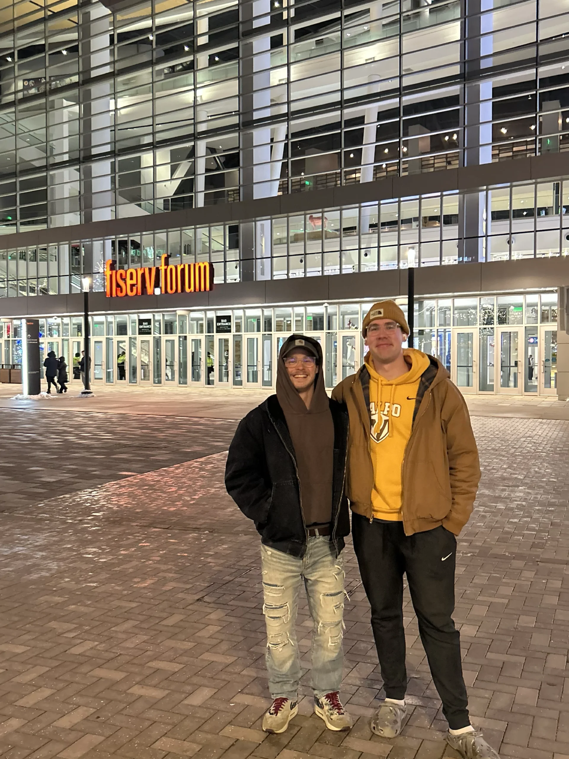 Parker Kelson and a friend stand smiling outside the brightly lit Fiserv Forum entrance at night, their faces illuminated like a beacon's reflection in the city.