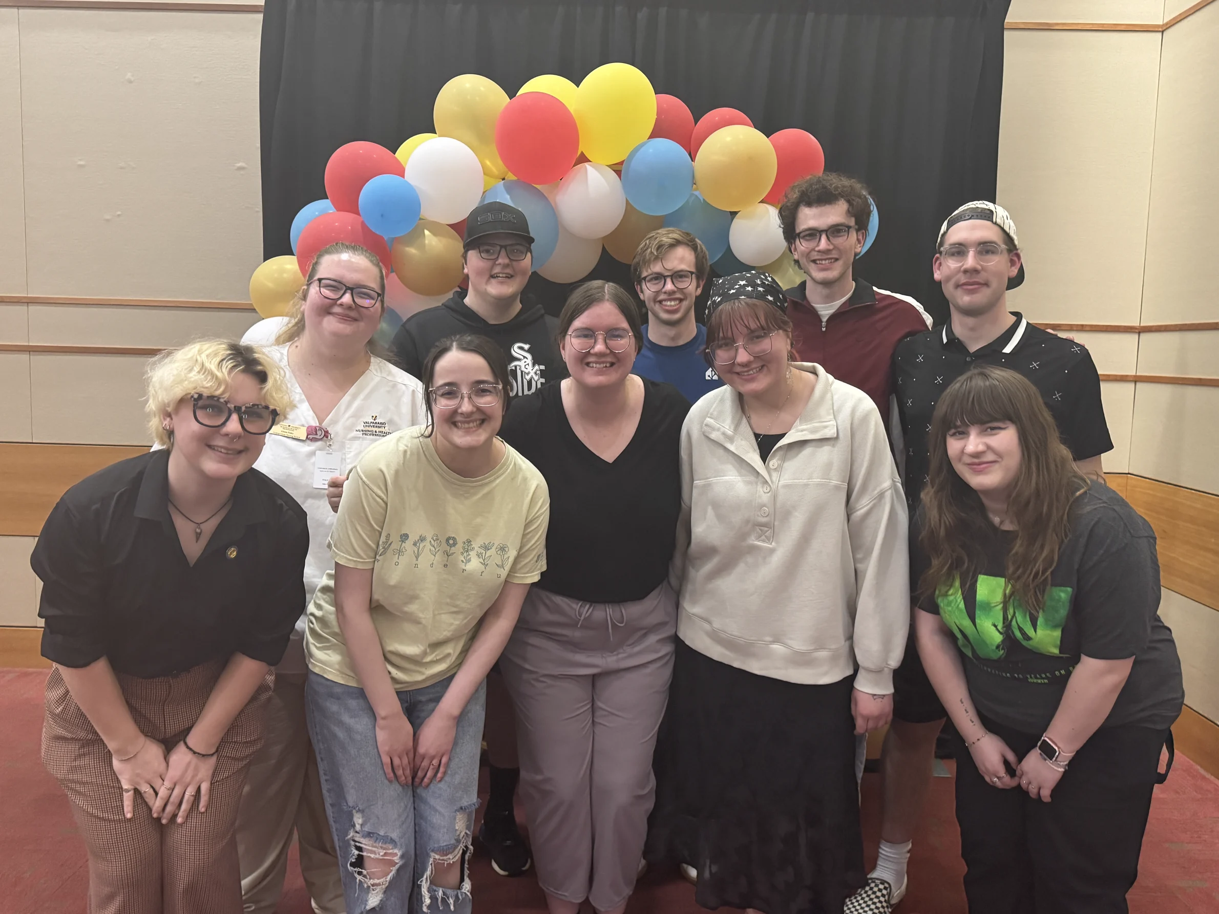 Ten people, including Parker Kelson, smile in front of a colorful balloon arch in a room with red carpet and tan walls, capturing the joyful spirit of Beacon's Reflection.
