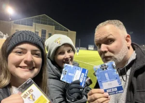 Three people in winter clothing, including AJ Stevens, hold media passes at a nighttime outdoor soccer field as the lights cast Beacon's Reflection across the pitch.